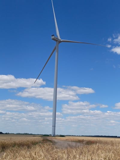 A lone wind turbine stands tall in a field of golden wheat under a bright blue sky dotted with white clouds. The turbine's blades are angled, with one pointing upwards and the other extending horizontally across the frame. A dirt path leads into the field, with some red flowers visible along its edge. The background shows a treeline and a flat horizon. The scene is set in Koshary, Ukraine, a rural location known for its agricultural landscape and wind farms.