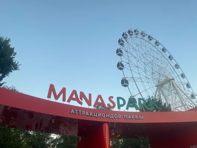 The image shows a Ferris wheel at an amusement park. The Ferris wheel is large and white, with many cabins attached to its spokes. The sky is clear and blue, suggesting it is daytime. In the foreground, there is a red structure with the words "MANASPARK" and "АТТРАКЦИОНДОР ПАРКИ" written on it. The word "MANASPARK" is in large red letters, with "PARK" in green letters. The reflection on the red structure shows blurry figures of people, implying they are walking past or entering the park. The overall scene evokes a sense of fun and recreation, typical of an amusement park setting.