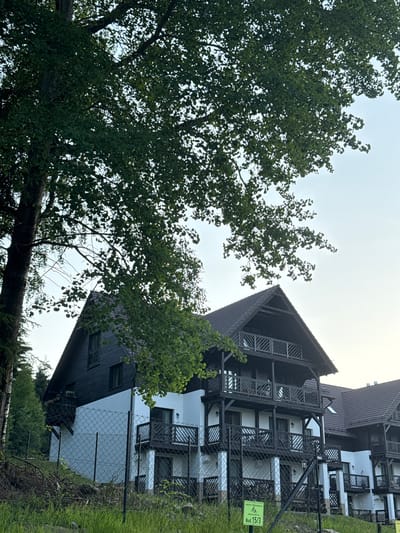 This image depicts a modern apartment building with a rustic design, located in Szklarska Poręba, Poland. The building features white lower levels and dark wooden upper levels, with multiple balconies adorned with wooden railings. The structure is nestled amidst lush greenery, with a large tree dominating the foreground, its leaves casting shadows on the building. A wire fence separates the property from a grassy area in front, where some branches and debris are visible. The sky is overcast, suggesting it might be a cloudy day. A small green sign with white lettering is attached to the fence, indicating "Bud. 15/3". The overall scene conveys a sense of a tranquil mountain resort or residential area.