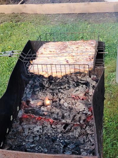 A close-up shot captures a barbecue grill in Soivolovskaya, Russia. The grill is filled with glowing embers and charcoal, with a metal rack holding a partially cooked food item, likely meat, positioned above the heat. The food appears to be light brown and has grill marks. The grill itself is a rectangular metal container, and it sits on a grassy area with scattered yellow wildflowers. In the background, a wooden plank and more grass are visible. The overall scene depicts an outdoor cooking setup.
