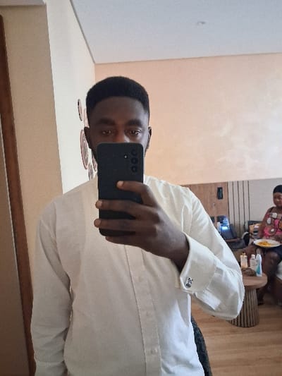 The image depicts an indoor scene within a room located in Takum, Nigeria. In the foreground, a dark-skinned male, with short dark hair and visible facial hair, is seen from the chest up, taking a mirror selfie. He holds a dark-colored smartphone, featuring a vertical triple-camera setup, in his right hand, which obscures his nose and mouth. He wears a light-colored (cream or off-white) long-sleeved collared shirt, with a silver-colored cufflink visible on his right wrist. In the background, to the right, a dark-skinned female is seated on what appears to be a light-colored couch or bed. She wears a multi-patterned top and dark bottoms, holding a white plate containing yellow food items. Adjacent to her, a small, round wooden side table with vertical slat-like legs holds several bottles and other small objects. Behind her, a piece of furniture with vertical dark slats is visible, along with an electronic device emitting a blue light. The room features light-colored walls and a wooden door frame is visible on the left.