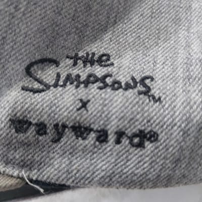 A close-up image depicts a gray, woven textile, exhibiting a distinct diagonal twill pattern consistent with denim fabric. Black embroidery is visible on the surface. The embroidered text, arranged vertically, reads "the SIMPSONS™ x wayward®". "The Simpsons™" is presented in a larger, stylized font, while "the" and "wayward®" are in smaller script. A small "x" serves as a separator between the two primary brand names. The fabric surface appears slightly worn, with minor loose fibers visible. There are no individuals, actions, or specific environmental features discernible in the frame beyond the textile and its embroidery. The precise location within Gimpo-si, South Korea, cannot be determined from this image.
