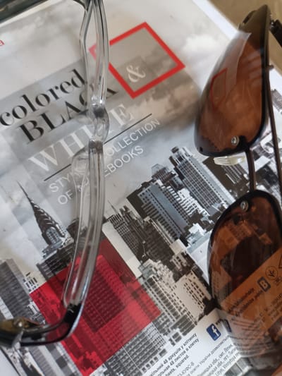 The image shows a pair of sunglasses resting on a notebook or magazine. The notebook/magazine features a black and white cityscape with a red geometric shape. Text is visible on the notebook/magazine in both English and Ukrainian. The sunglasses have brown lenses and clear frames. One lens has text reflected on it, which appears to be website addresses in Cyrillic script. A business card is partially visible at the bottom right, with a Facebook logo and contact information in Ukrainian, listing an address on Khmelnytskoho St. in Horokhiv, Ukraine. The overall lighting is bright, suggesting an indoor setting with overhead lighting or natural light from a window. There are no people visible in the image.