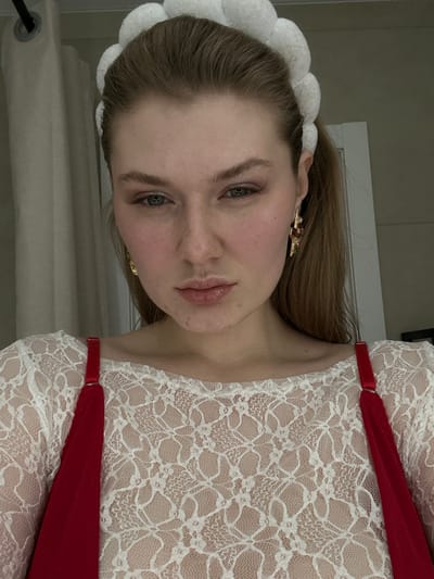 A woman with fair skin, blue eyes, and light brown hair looks directly at the camera. She wears a white, lacy top with red straps and a large, white, fluffy headband. Gold earrings are visible on both ears. The background is blurred, but a white doorframe is visible. The image is slightly rotated, giving it a tilted perspective.