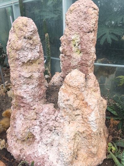 The image displays a detailed view of three large, irregularly shaped, textured formations, resembling termite mounds or porous rock structures, within an indoor horticultural display or greenhouse facility in Munyati, Zimbabwe. The structures exhibit a predominant reddish-brown and off-white coloration, with patches of pale yellow and darker soil-like material. The two most prominent formations stand vertically, flanking a smaller, central structure that emerges from the ground. All formations have a rough, uneven, and somewhat pitted surface texture. In the background, partially obscured by the formations, are glass panels and various green plants, including slender cacti and broader-leafed foliage, indicative of a controlled growing environment. Additional smaller cacti and succulents are visible on the ground level around the base of the formations. No human figures are present, and no actions or interactions are depicted. The scene is static, focusing on the artificial landscape and plant life.