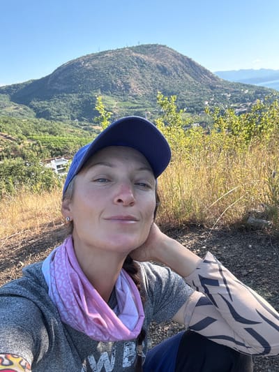 A woman with fair skin, wearing a blue baseball cap and a grey hoodie, takes a selfie outdoors. She has a pink and white patterned neck gaiter around her neck. Her right arm is resting on her head, and she is wearing a tan arm sleeve with black tiger stripes. In the background, a large, tree-covered hill dominates the landscape under a clear blue sky. The hill slopes down to reveal a town with white buildings nestled amongst greenery and rows of what appear to be vineyards. Further in the distance, a body of water and other hills can be seen. The foreground consists of dry grass and rocky, earthy terrain.