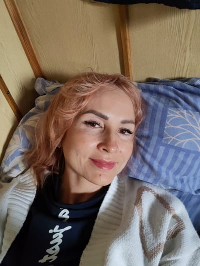 A young woman with short, pinkish-blonde hair and makeup is lying in bed. She is wearing a dark blue t-shirt with white lettering and a white cardigan with a tan geometric pattern. The bed has a blue and white striped pillow with a leaf-like pattern. The background appears to be the interior of a dwelling, with wooden walls and beams visible. The image is a close-up, selfie-style shot. The location is a bedroom in Vytegra, Russia.