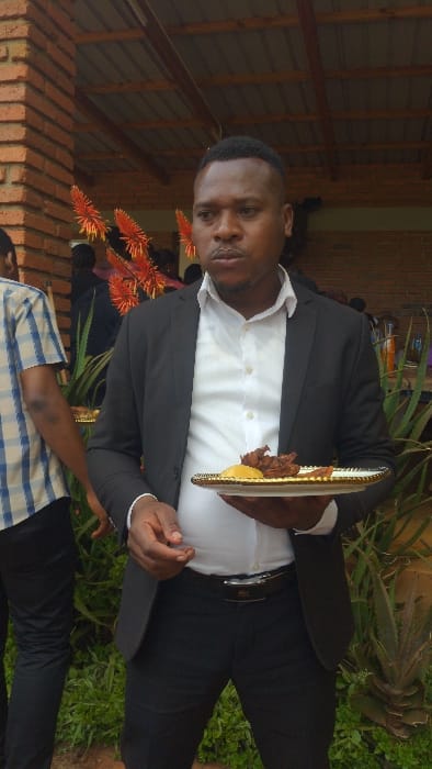 A man in a black suit and white shirt holds a plate with food. He stands near orange flowers and greenery, with a brick structure visible behind him. Another person, wearing a blue and white plaid shirt, is partially visible in the lower part of the image. The setting appears to be outdoors.