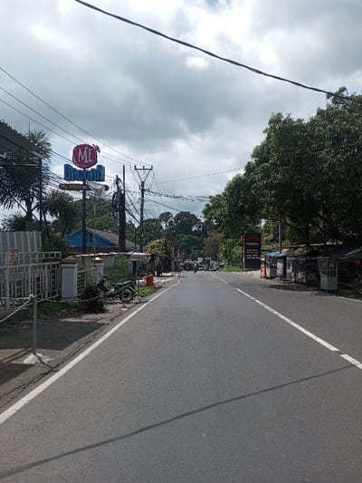 The image displays an upward-sloping, two-lane asphalt road in Gubbisamamidi, India, under an overcast sky. The road is marked with a solid white line on its right edge.
On the left side of the road, in the foreground, a white metal fence with a chain barrier delineates a property. Behind it, a black motorcycle is parked on a dirt shoulder next to a small, light-colored structure. Further back, a vibrant blue building is partially visible. A prominent sign featuring a red oval with "ME" and blue text "GACOAN" is mounted on a pole near multiple utility poles densely covered with tangled wires. Palm trees and other green foliage are visible behind these structures.
On the right side of the road, a row of small, informal roadside stalls or kiosks with varying roof materials (corrugated metal, tarps) line the shoulder. Behind these stalls, a large, dark rectangular sign displays text, with "PENJURIA" visible at the top. A dense canopy of large green trees extends along this side of the road.
In the distance, along the ascending road, several indistinct vehicles, possibly motorcycles and a car, are visible. The scene depicts a semi-urban or rural thoroughfare with commercial activity. No specific individuals are performing discernible actions in the immediate foreground.