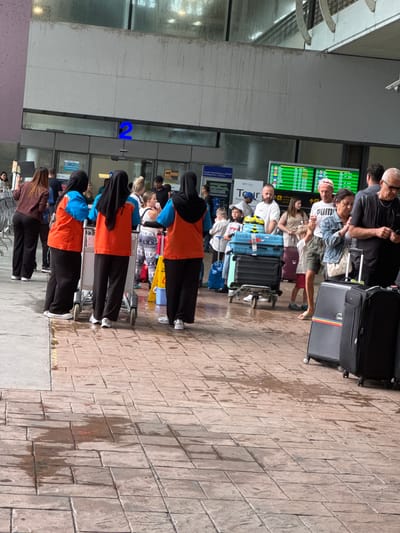 The image depicts an outdoor scene at Phuket International Airport in Tambon Mai Khao, Thailand, likely in the arrivals or departure area given the presence of luggage and people. The ground is paved with interlocking bricks and shows wet patches, suggesting recent rain or cleaning. A blue sign with the number "2" is visible above an entrance.

Several individuals are present. Three people wearing orange vests and black headscarves are pushing luggage carts. One person is wearing a brown jacket and black pants, facing away from the camera. Others in the background are also holding or near luggage.

Various suitcases are visible, including a large blue suitcase stacked with a smaller black suitcase on a luggage cart. Other dark-colored suitcases are also present. In the background, a digital display board shows flight information. The overall atmosphere appears to be one of travel and transit.