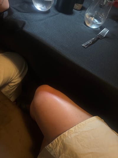 The scene is located in Spanish Town, British Virgin Islands. The image depicts a close-up, downward-angled view of a dining setting in a low-light environment. In the foreground, the upper portion of a bare, tanned human leg, wearing light-colored shorts or a skirt, occupies the lower right quadrant. To the left, a portion of another person's leg or light-colored garment is partially visible. The upper half of the frame is dominated by a black tablecloth. On the tablecloth, from left to right, a clear glass containing a cold liquid with condensation, a dark cylindrical object (possibly a condiment shaker), and a second clear glass with a cold liquid are arranged. A silver four-tined fork rests diagonally on the black tablecloth in the upper right section of the image. The lighting appears localized, illuminating the visible leg and table items against a darker background.