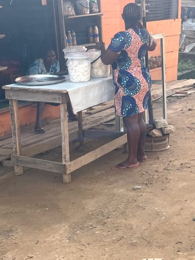 In Ijegun, Nigeria, an image captures a woman standing at a wooden table, likely a street vendor setup. The table displays various goods, including items in transparent jars and other packaged products. The woman wears a patterned dress and sandals. The surrounding environment includes a dusty ground surface and other buildings or stalls. Another person is visible behind the table. The overall setting suggests a local market or commercial area.