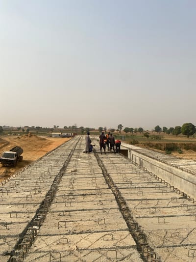 The image displays a construction site for an elevated concrete structure, likely a bridge or roadway, in Wuciciri, Nigeria. The scene is observed under a pale, hazy sky. In the foreground and extending into the midground, the concrete structure is visible, composed of numerous rectangular pre-cast concrete slabs. Exposed U-shaped rebar loops project upwards from the slabs, and continuous linear rebar cages are present within longitudinal channels between the slab rows. Seven adult males are positioned on the concrete structure in the midground. One male, wearing a light-colored full-length robe, stands to the left of a cluster of six other males. This cluster is gathered towards the right edge of the current concrete section, appearing to observe or engage in an activity at the construction interface. To the left of the concrete structure, a white tanker truck with a dark grey cylindrical tank is stationary on exposed reddish-brown earth. Further left, mounds of excavated earth and several distant temporary buildings are discernible. The right side of the scene extends into undeveloped terrain characterized by scattered trees and low vegetation under a hazy horizon.