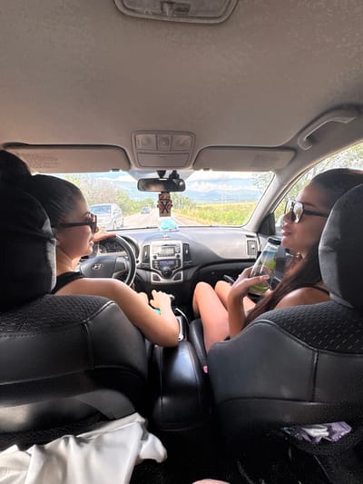 The image depicts the interior of a Hyundai vehicle, observed from a rear passenger seat, in motion on a road in Marino Pole, Bulgaria. Two females are present in the front seats. The driver, seated on the left, has dark hair pulled back, wears sunglasses, and is operating the steering wheel, looking forward and slightly right. Her right hand is positioned near the gear shifter. The front passenger, seated on the right, has long dark hair, wears large sunglasses, and is holding a clear plastic bottle containing green liquid to her mouth, appearing to be drinking. Her legs are extended, and a white charging cable is visible near her seat.

The car's interior includes a dark dashboard, a Hyundai-branded steering wheel, and a center console featuring a stereo unit displaying "10/10/10," air vents, and climate controls. A rearview mirror is centrally positioned, from which an "Areon" branded air freshener and a small blue plush toy are suspended. The light-colored headliner exhibits minor discoloration. Dark upholstered seats are present. On the floor behind the driver's seat, a crumpled white garment is visible. Behind the passenger's seat, a small purple packaged item rests on the floor.

Through the windshield, a two-lane asphalt road is visible with another silver vehicle ahead of the subject car. The exterior environment consists of green vegetation lining the road and distant mountains under a partly cloudy sky, indicating a rural or semi-rural setting.