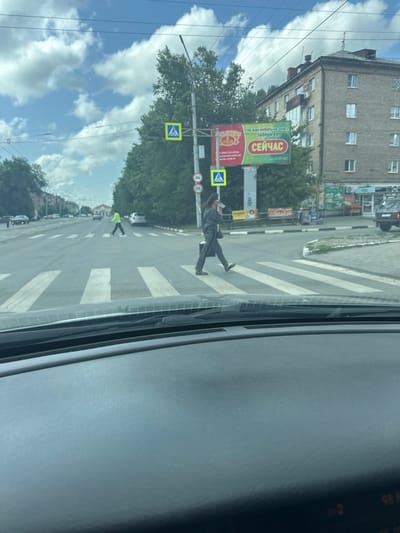 The image captures a street scene in Orsk, Russia, on a cloudy day. In the foreground, the dashboard of a car is visible, partially obscuring the view ahead. The main focus is a crosswalk on a multi-lane road. Two pedestrians are crossing: a person in a bright yellow jacket is in the left lane, and another person in a dark outfit is in the right lane. A yellow pedestrian crossing sign is positioned on the left side of the road. In the background, a multi-story brick building stands, with trees and a large billboard to its left. Several cars are present on the road and parked on the side. The sky is filled with white, puffy clouds against a light blue backdrop.