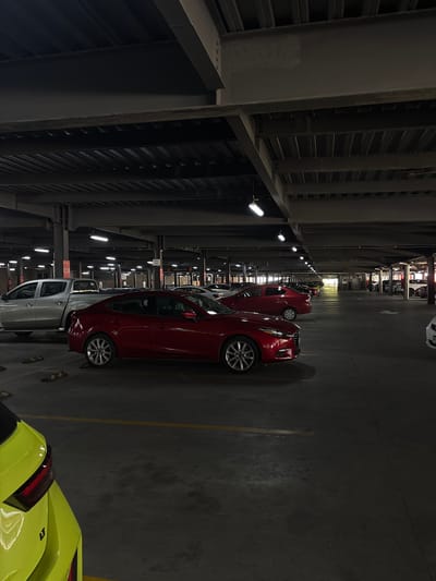 The image shows a parking garage in what appears to be San Pedro Cholula, Mexico. The location is indicated by signage in Spanish. Several cars are parked in a row. A red hatchback is prominently displayed, along with parts of a silver pickup truck and a bright yellow sports car. The garage is illuminated by fluorescent lights attached to the structure of the building. The garage is constructed of steel. The lighting is dim and creates deep shadows.