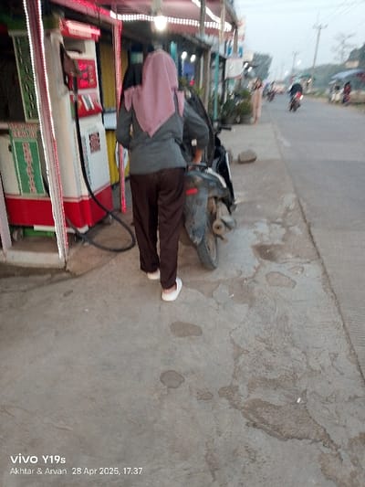 The image shows a woman in a grey shirt and brown pants wearing a pink headscarf standing next to a motorcycle. She is in the process of preparing the motorcycle for travel. The motorcycle is facing right and is parked in front of a gas station with red and white paint. The woman's left foot is visible, and she is facing left. The scene takes place in the town of Kecamatan Kroya, Indonesia. The woman is on a sidewalk made of concrete. The sidewalk is cracked and uneven. The pavement is stained with a water-like substance, which is either the result of a spilled drink or a rainstorm. The gas pump is at the corner of a building. The gas pump is visible in the background of the image. The motorcycle is parked in the middle of the sidewalk. The woman's right hand can be seen touching the motorcycle. The woman is standing near the gas pump. It is a cloudy evening. The scene is taking place in an urban environment. The ground is concrete and cracked in places. There are trees in the background of the image. There is an empty street with a motorbike in the distance, and there is a building behind the woman. The image is taken at an angle looking down on the scene. The image is blurry.