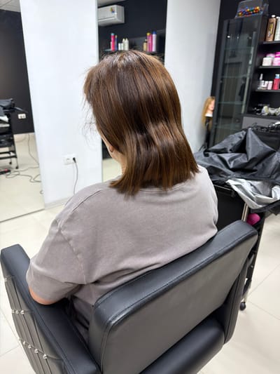 The image shows the back of a person with medium-length brown hair sitting in a black leather salon chair. The person is wearing a light gray t-shirt. The salon appears to be modern and clean, with white walls and tiled flooring. In the background, there is a mirrored wall, shelves stocked with hair products, and a mannequin with blonde hair. There are also some professional salon equipment visible, such as a rolling cart and a black tarp. The lighting is bright and even, suggesting an indoor setting. The location is identified as a salon in Timashevsk, Russia.