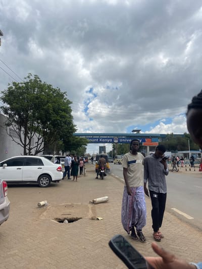 This image depicts an urban street scene in Thika, Kenya, specifically identifiable by the "Mount Kenya University" archway, placing it within the vicinity of Hospital Ward. The sky is largely overcast with heavy grey clouds, interspersed with patches of blue sky.
In the immediate foreground, the top of a person's head with braided hair and a hand holding a black smartphone are partially visible on the right, indicating the photographer's position.
The scene focuses on a reddish-brown paved sidewalk (composed of pavers) and an adjacent asphalt road. On the sidewalk in the mid-ground, two young adult males are walking right-to-left from the viewer's perspective. The taller male on the left wears a light-colored t-shirt with a horizontal dark stripe and a patterned light purple sarong-like garment, and brown sandals; he is looking towards the camera. The male next to him wears a dark patterned long-sleeved shirt and dark trousers, and light-colored flip-flops; he is looking down at a smartphone he holds.
The sidewalk features a safety hazard: a missing manhole cover, revealing a dark hole with debris inside. Two white cylindrical objects, resembling discarded gas cylinders or construction pipes, lie on the sidewalk near the open manhole.
To the left of the sidewalk, a white four-door sedan is partially parked, with its trunk open. A group of approximately five to seven pedestrians is visible standing or walking near this vehicle. A large green tree with dense foliage stands behind the parked cars.
The two-lane road carries light traffic, including a yellow auto-rickshaw (tuk-tuk) and a motorcycle with a rider. Across the road, additional pedestrians are visible on another sidewalk. An elevated archway, prominently displaying "Mount Kenya University" in blue and orange lettering with university logos, spans the road. Overhead utility lines are visible against the sky.