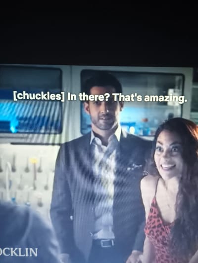 The image depicts two individuals, a man and a woman, within an indoor setting that resembles a scientific laboratory. The man, positioned centrally, has dark hair and a beard, wearing a dark suit jacket over a patterned light-colored shirt, and displays a slight smile while looking forward. To his right, the woman, with long dark wavy hair, wears a red and black animal-print dress and exhibits a wide, excited smile, looking towards the left. The on-screen text reads "[chuckles] In there? That's amazing." In the background, to the left, various pieces of laboratory glassware and tools are mounted on a white panel. Behind the man, illuminated panels with a blue glow are visible. The shoulder of a third person is partially visible in the bottom-left corner, with the text "OCKLIN" below. The scene is located in the city of Panicheri, Bulgaria.