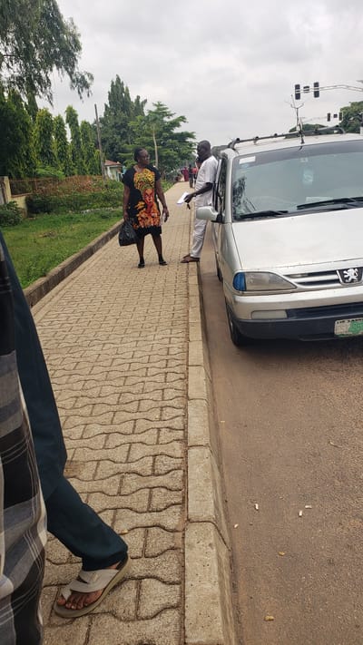 In Unguwar Shanu, Nigeria, on an overcast day, a silver sedan is parked on the right side of a road, its front passenger door ajar. A man in a white tunic and trousers stands next to the open door, facing a woman who is also standing. The woman wears a black dress with a vibrant, graphic print of a woman's face and upper body. She carries a dark bag in her left hand. Both individuals appear to be engaged in conversation. The sidewalk, paved with light brown interlocking bricks, runs parallel to the road. Beyond the sidewalk, a line of well-maintained, conical trees borders a grassy area. In the background, traffic lights are visible above the trees. The foreground shows the lower legs and feet of another individual wearing dark trousers and sandals, walking on the sidewalk. The scene is captured from a slightly elevated perspective, looking down and across the sidewalk towards the vehicle and the two conversing individuals.