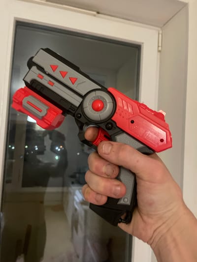 A person's hand is holding a red and gray toy gun in front of a window. The gun has a red trigger and a gray grip. There are red triangle markings on the top of the gun. The window appears to be a sliding glass door, with reflections of lights visible in the glass. The background outside the window is dark, suggesting it is nighttime. The scene is indoors, with the window frame and a wall visible. The location is Laishevo, Russia.