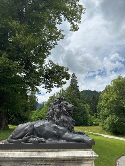 The image depicts a bronze lion sculpture in the foreground, seated on a pedestal in a park-like setting. The sculpture is dark and detailed, with a prominent mane. Behind the sculpture, lush green trees and rolling hills dominate the landscape. A gravel path winds through the grassy area, leading into the dense forest. The sky is cloudy with patches of blue sky visible. In the distance, mountains are visible, shrouded in mist. The scene is peaceful and serene, suggesting a natural or historical site. Based on the surroundings and the architecture, this location is likely within the grounds of Ettal Abbey in Ettal, Germany, a Benedictine monastery renowned for its baroque architecture and picturesque setting. There are no people visible in the image.