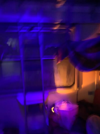 A dimly lit space with a metal grid in the foreground is illuminated by blue light. Behind the grid, a train window reflects an indistinct exterior scene bathed in golden light. A translucent plastic bag is affixed to the wall. The scene is out of focus, rendering details obscure. The composition includes the back of a person, barely discernible in the background.