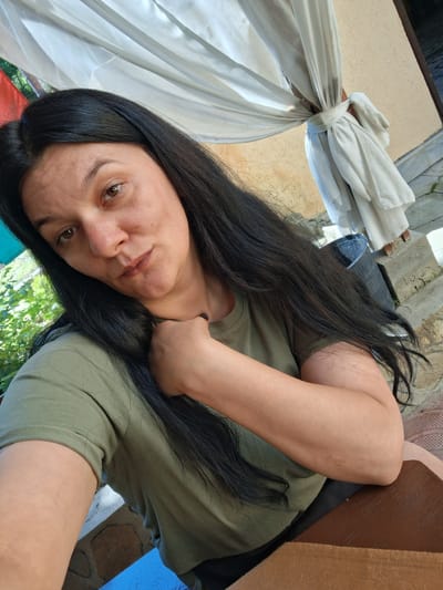 The image shows a woman with long, dark hair and brown eyes, wearing a green t-shirt. She is seated at a wooden table, with her right arm resting on her chest. The background features a beige wall, white drapery, greenery, and a black bucket. The image was taken outdoors in the city of Marchevo, Bulgaria.
