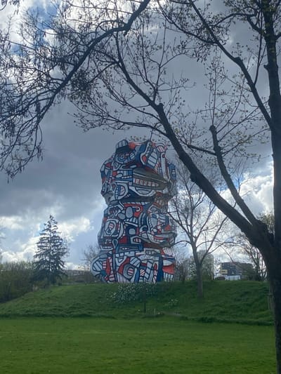 A large, multi-tiered abstract sculpture, identified as the Tour aux figures by Jean Dubuffet, is centrally positioned on a grassy mound within Parc Henri Barbusse in Issy-les-Moulineaux, France. The sculpture features an irregular, organic form covered in a dense pattern of interlocking, stylized shapes outlined in black and filled with red, blue, and white.

The immediate foreground consists of a vibrant green lawn. Clusters of small white flowers are visible at the base of the mound, to the right of the sculpture. To the left of the sculpture, a tall, conical evergreen tree stands on the hillside. Distant urban buildings are faintly discernible on the horizon line in the background. Bare tree branches, some with nascent buds, frame the upper portion of the image, silhouetted against a sky showing a mix of dark clouds and brighter patches. No individuals or active subjects are present in the scene.