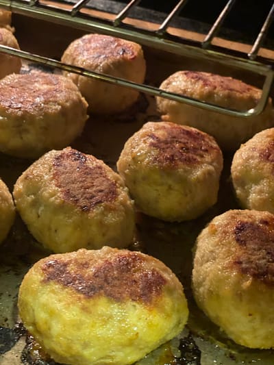 The image shows a close-up of several cooked, round patties, possibly meatballs or croquettes, arranged on a baking sheet. The patties have a golden-brown, slightly browned surface with some darker, crispy areas. They appear to be resting on a dark, possibly greasy surface, with hints of a metal rack above them. The lighting suggests they might be under a broiler or in an oven. The scene depicts food being prepared or cooked. There is no indication of the city of La Línea de la Concepción, Spain, or any elements that would suggest this specific location. The image is purely a still life of food.