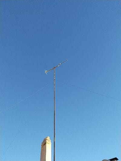 An antenna stands tall against a bright blue sky. The antenna is composed of a central pole with horizontal metal bars extending outwards. Thin wires stretch diagonally from the antenna, nearly invisible against the sky. Below the antenna, a light-colored structure, possibly a chimney, juts out from a roofline. The bottom of the image suggests the top of a building or house.