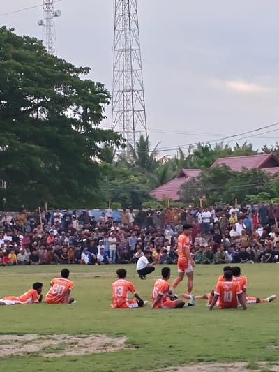This media file captures a football match taking place in Desa Meunasah Blang, Indonesia. The scene is set on a grassy field with a large crowd of spectators gathered to watch. In the background, tall telecommunication towers dominate the sky, and lush green trees and buildings with red roofs are visible. The time of day appears to be late afternoon or early evening, as indicated by the soft, diffused light and the hazy sky, which might suggest cloudy weather.
The primary subjects are two groups of people: the football players on the field and the spectators. The players, all wearing bright orange jerseys, are mostly seated on the grass, some with their backs to the camera. One player stands in the center of the field, looking towards the spectators. The spectators are a diverse group, many dressed in casual attire, with some wearing headscarves. Their presence suggests a community event or local competition.
The overall mood seems to be one of casual observation, with the focus on the sporting event and the communal gathering. The image conveys a sense of local life and leisure activities in a semi-rural Indonesian setting.
Visible text on the jerseys includes "ARM FC" and numbers such as "30", "13", and "10", indicating the team affiliation and individual player identification. The telecommunication towers are prominent objects that provide a distinctive landmark to the location.