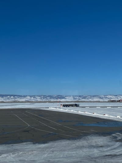 The image shows a bright blue sky dominating the left half of the frame. On the right is an outdoor, snow-covered area with mountains in the distance. A dark-colored train is visible on a straight track near the mountain range. The ground, partially covered in snow and ice, shows marked lines and dark patches of what appears to be a road or runway.