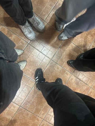 The image shows a view of the lower bodies of five people standing in a circle on a tiled floor. Each person is wearing different footwear: grey suede shoes, black leather shoes, black Adidas sneakers, white sneakers, and grey suede cowboy boots. The people are wearing trousers of different colors, including grey, black, and denim. The tiled floor appears to be inside a building. The image location is San Juan Cuautlancingo, Mexico.