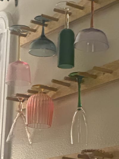The image displays an interior scene within a building in the city of New Orleans, United States. A light-colored wooden rack is affixed to an off-white, textured wall. This rack is constructed with horizontal wooden pegs designed to suspend stemware upside down.
Six distinct glasses are currently hung from the rack. In the upper row, from left to right, these include a pink ribbed wine glass with a gold-toned stem, a blue-grey stemmed cocktail glass, a dark green cylindrical glass resembling a repurposed bottle, and a light purple/clear wide-rimmed stemmed glass.
In the lower row, also from left to right, are a clear ribbed stemmed cocktail glass, a pink ribbed wine glass identical to the upper-left, and a clear champagne flute featuring a green stem.
To the far left, a narrow vertical segment of a white architectural feature, consistent with a door or window frame, is visible. In the lower right corner of the frame, a portion of another wooden surface is present, upon which several small, irregular brown objects are arranged. No individuals are depicted in the scene.