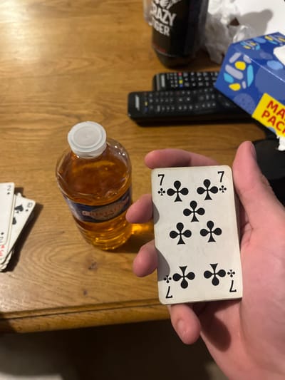 The scene depicts an indoor setting on a light-brown wooden table in Amiens, France. A human hand holds a white playing card, prominently displaying the "7 of Clubs." To the left of the hand, a transparent plastic bottle with a white cap contains an amber-colored liquid, partially revealing a "CRISTALINE" label. Further to the left, a partial stack of additional playing cards, including a '2 of Spades' and '10 of Spades', is visible. In the upper right background, two black remote controls are positioned next to a blue and yellow "MAXI PACK" tissue box, alongside a crumpled white tissue. A dark beverage container with "CRAZY TIGER" text is also partially visible in the upper background.
