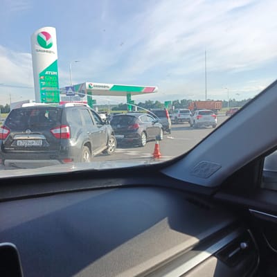 In Bulgakovo, Russia, a queue of vehicles is forming at a Bashneft gas station under a clear, cloudy sky. The gas station features a prominent, tall white sign with the Bashneft logo, displaying fuel prices for ATUM, 95, 92, and AI-92 octane fuels. The canopy of the gas station is visible, with its green and pink trim. The foreground is dominated by the interior of a vehicle, showing a dashboard with a textured surface and a speaker grille. A black Nissan Terrano is positioned in the immediate foreground, its rear end facing the camera, displaying a license plate "B709HP702". Other vehicles, including a dark-colored hatchback, a white SUV, and a truck, are visible further back in the queue. The scene suggests a typical day at a Russian gas station, with cars waiting for service.