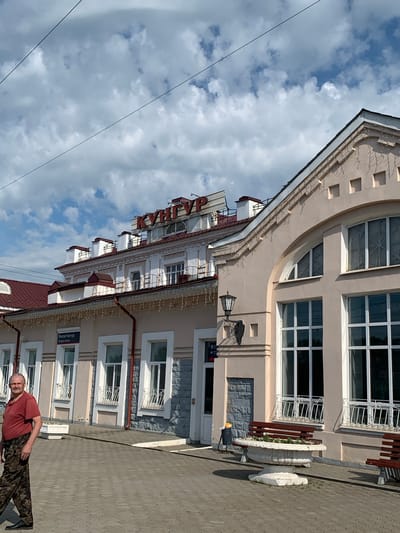 The image shows a man lying on the ground in front of a building in Kungur, Russia. The man is wearing a red t-shirt and camouflage pants. He appears to be relaxed and is looking at the camera. The building behind him is light beige with several windows and decorative trim. A sign with Cyrillic text is visible on the building. The sky is partly cloudy. The overall impression is of a casual outdoor scene on a bright day.