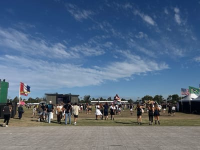 The image depicts an expansive outdoor event scene in a large field within San Isidro, Argentina, under a clear blue sky with scattered cirrus clouds. The foreground consists of grey, interlocking ground panels. Numerous individuals, predominantly young adults in casual attire, populate the vast grassy area, walking and standing.
In the mid-distance, two large outdoor stages, each featuring prominent video screens, are visible. To the left of the scene, a vertical French flag (blue, white, red) flies from a flagpole, positioned next to a smaller red flag and a tall green structure. To the right, three flags fly from individual poles: the United States flag, a green flag (possibly Brazilian), and a third flag partially obscured. These flags are situated near white canopy tents. A large, light-colored inflatable or statue, resembling a stylized animal or character, stands erect in the mid-right portion of the field. A line of trees forms the distant horizon, bordering the event grounds.