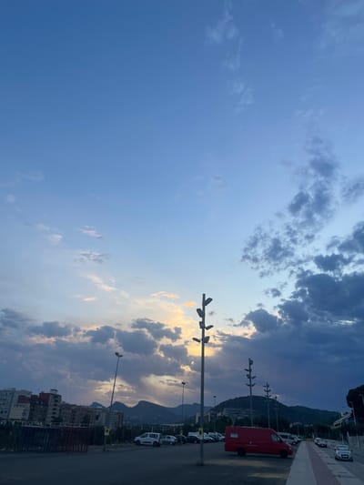 This is a wide-angle, horizontal, outdoor photograph taken at dusk in the city of Sagunto, Spain. The sky dominates the upper two-thirds of the frame and is filled with a mixture of light blue and dark, dramatic clouds. The sun is setting behind the clouds on the left side of the image, casting a warm, yellowish glow. Several tall, multi-headed streetlights are visible, standing in a row across the mid-ground of the image.
The lower portion of the image shows a paved area, likely a parking lot or street, with several parked vehicles. A bright red van is parked in the center-left of the frame. Beyond the parking area, a range of hills and mountains forms the backdrop, with buildings nestled on their slopes. Some multi-story residential buildings are visible in the left mid-ground. A sidewalk with red and white markings runs along the right side of the paved area. The overall impression is that of an urban landscape at the end of the day, with the natural beauty of the sky and mountains contrasting with the man-made structures. No people are visible in the photograph.