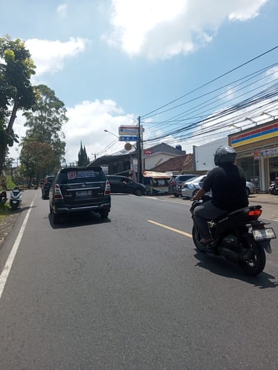 The image shows a road in an urban area, potentially in Asia based on the store signs. A black car with license plate "F 1443 KO" is visible, followed by other cars, while a person on a motorcycle with license plate "F 5523 WAX" is driving on the right side of the road. An "Indomaret" store and an ATM are on the right side of the road. The sky is blue with clouds. The foreground consists of the road's asphalt surface.
