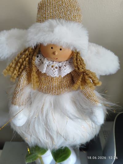 A close-up, front-facing shot shows a decorative angel figurine. The angel has a smooth, light-brown, round face with simple painted features: small black eyes, a small red smile, and a light-brown nose. It wears a knitted hat with gold and white threading, and a fluffy white trim that extends to form large wings. The angel's hair is made of thick, golden-brown yarn, styled into two braids that hang down its sides. Its body is covered in a white, fluffy material, reminiscent of snow or soft fur, with thin gold strands woven throughout. The angel's torso is adorned with a knitted, gold and white striped sweater and a white lace collar. The overall impression is festive and perhaps Christmassy. The background is a plain, light-colored wall. In the bottom right corner of the image, a timestamp is visible: "18.06.2026 12:26". There are no clear indicators of weather or time of day beyond the timestamp, and the location context is Northampton, United Kingdom, although this is not visually evident from the image itself. The figurine appears to be an inanimate object, so no activity or emotions are depicted.