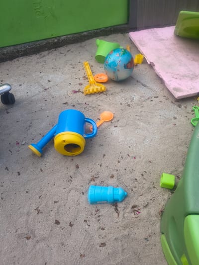 The image shows a collection of children's toys scattered on a concrete and sandy surface. In the upper left, a pink rectangular object, possibly a mat or board, is visible. Next to it are parts of green plastic playground equipment. Scattered on the ground are various toys including a blue and yellow watering can, a small globe with a picture of a face, a yellow sand shovel with a star-shaped mold, and a few orange sand molds. A bright blue cylindrical object, possibly a bottle or container, is positioned on the sand. The ground is a mixture of concrete and sand, with some dried leaves and debris scattered around. In the lower left corner, a green surface with the word "BIG" and a silhouette of an animal is visible, suggesting it's part of a larger toy or play structure. The scene appears to be an outdoor play area, likely a backyard or playground, given the presence of sand and children's toys. The lighting suggests it's daytime, and the overall impression is one of playful disarray. No people are visible in the image.