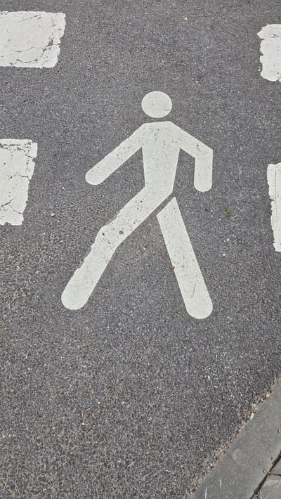 The image shows a pedestrian crossing symbol painted in white on asphalt in Ūlupji, Latvia. The symbol depicts a stylized human figure walking. The asphalt is dark grey and appears to be slightly worn, with some small pebbles visible. There are also white painted stripes of the pedestrian crossing, some of which are cracked and faded. To the right of the pedestrian symbol, a concrete curb is visible, indicating the edge of the road. The overall scene suggests an urban or semi-urban environment where pedestrian safety is marked on the roadway.