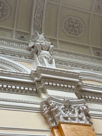 The image shows an ornate ceiling interior with intricate plasterwork, suggesting it is a historical building. A white stucco sculpture of a cherubic figure with eagle wings is prominently displayed, supporting a decorative element. The ceiling itself is coffered with geometric patterns and floral motifs. The overall aesthetic is classical and opulent. This ceiling is located in the Grand Palace of Gatchina, Russia, a former imperial residence.