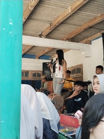 The image shows a live performance in Jenawi, Indonesia. A woman with long black hair stands on a makeshift stage holding a microphone. She is wearing a white sleeveless top with designs and gray pants. Behind her, a band is set up with a drummer in a yellow shirt, a keyboardist with blonde hair, and percussionists. There are also drums and other instruments in the scene. In the foreground, there are audience members, including one wearing a hijab and another holding a small electronic device. The location appears to be an open-air area with a corrugated metal roof supported by wooden beams and a turquoise pole on the left side. There are windows behind the stage. The lighting suggests it is daytime.