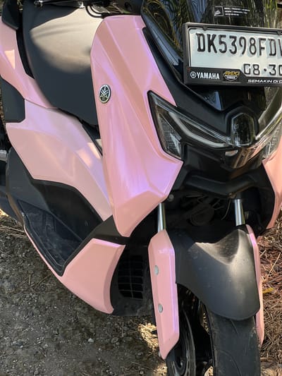 A close-up, high-angle shot shows the front of a pink motorcycle parked on a dirt and gravel surface, suggesting a rural or unpaved area, possibly in or near Kerambitan, Indonesia. The motorcycle has a black seat and black accents on the body. A license plate with the characters "DK5398FDW" and "08-30" is visible on the front. Yamaha logos are present on the motorcycle and on stickers below the license plate. The front wheel and fender are visible, as are parts of the headlight assembly. The scene is outdoors, with dry grass and twigs on the ground, indicating a sunny day.