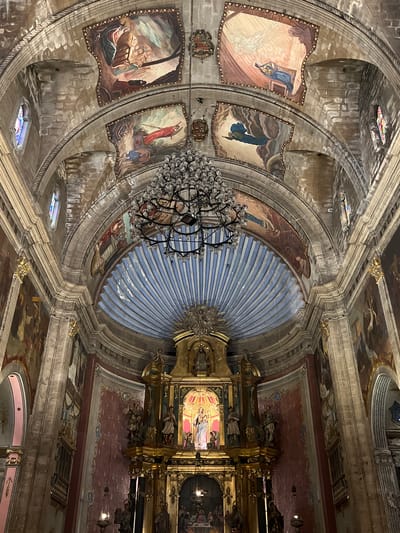 The image depicts the interior of a grand church, likely a cathedral or a significant parish church, with an ornate altar at the center. The ceiling is adorned with intricate frescoes depicting religious scenes and figures, framed by stone arches. A large, multi-tiered chandelier hangs from the center of the ceiling, casting a warm light on the scene. The walls are also decorated with large paintings and religious iconography. The altar itself is a complex structure, featuring multiple statues and gilded ornamentation. The overall impression is one of historical and religious grandeur, with a focus on elaborate artwork and architecture. The location is the Church of Our Lady of the Angels in Pollença, Spain.