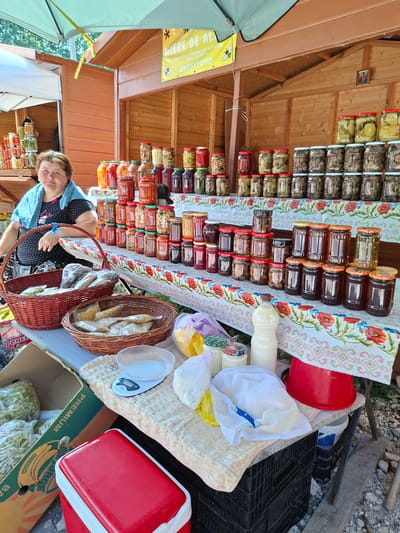 The image depicts an outdoor market stall in Scoreni, Moldova, where a woman is selling jars of preserved food and other goods. The stall is constructed of wood, with a green awning providing shade. A sign at the top reads "MIERE DE AL."
The woman, who appears to be in her 50s or 60s, is wearing a black dress with white polka dots and a light blue scarf around her neck. She is seated and looking towards the camera. In front of her are several wicker baskets containing bagged goods, possibly herbs or dried foods.
The main focus of the stall is the extensive display of glass jars filled with various preserved items. These include what appear to be pickles, jams, honey, and other pickled vegetables. The jars are neatly arranged on shelves and the table.
On the table in front of the jars are additional items: a digital scale, several bags of white produce (possibly cheese or dough), a bottle of white liquid (likely milk or yogurt), and a red bucket. To the left of the table, a cardboard box labeled "PREMIUM" and featuring a banana illustration contains more bagged green produce. A red and white cooler is also visible to the left.
The ground beneath the stall is covered with gravel and small stones, typical of an outdoor market setting. The overall impression is a local vendor selling homemade or locally sourced food products.